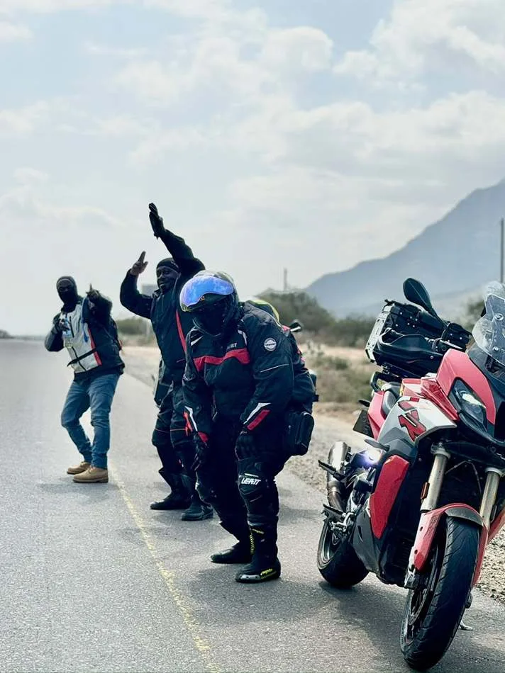 Riders saluting on the roadside beside a red motorcycle with mountains behind